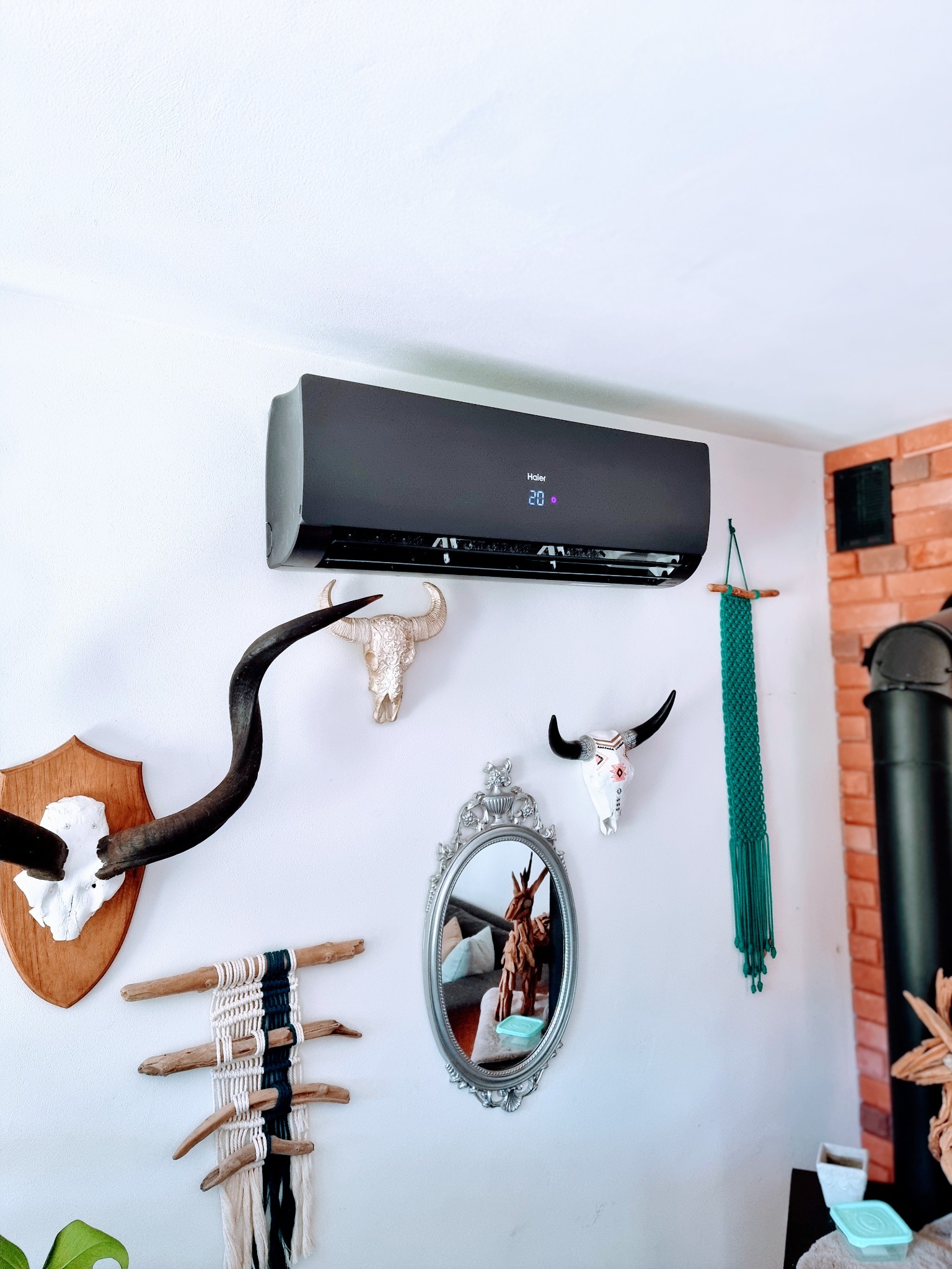 Czarny klimatyzator Haier zamontowany na białej ścianie w pomieszczeniu z dekoracjami w stylu boho: czaszki zwierząt, lustro w ozdobnej ramie i makrama. Widoczny fragment kominka z cegły.