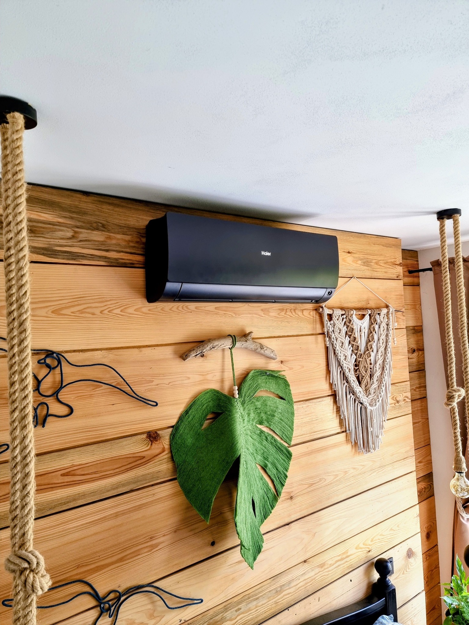 Czarny klimatyzator Haier zamontowany na drewnianej ścianie w pokoju, z dekoracjami w stylu boho: makrama i filcowy liść monstery.