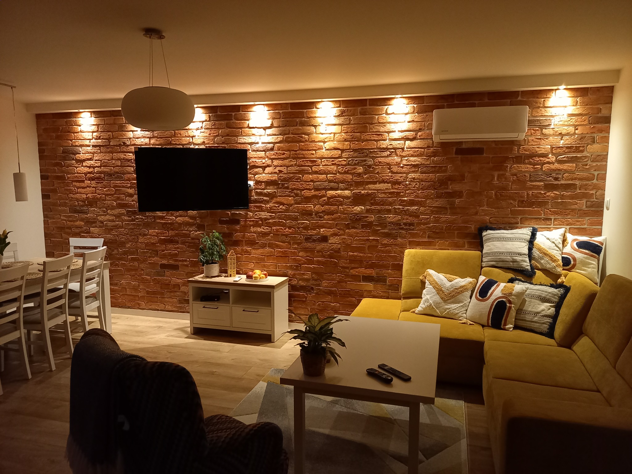 Salon z ceglaną ścianą, zamontowaną klimatyzacją, żółtą sofą i drewnianą podłogą w ciepłym oświetleniu.