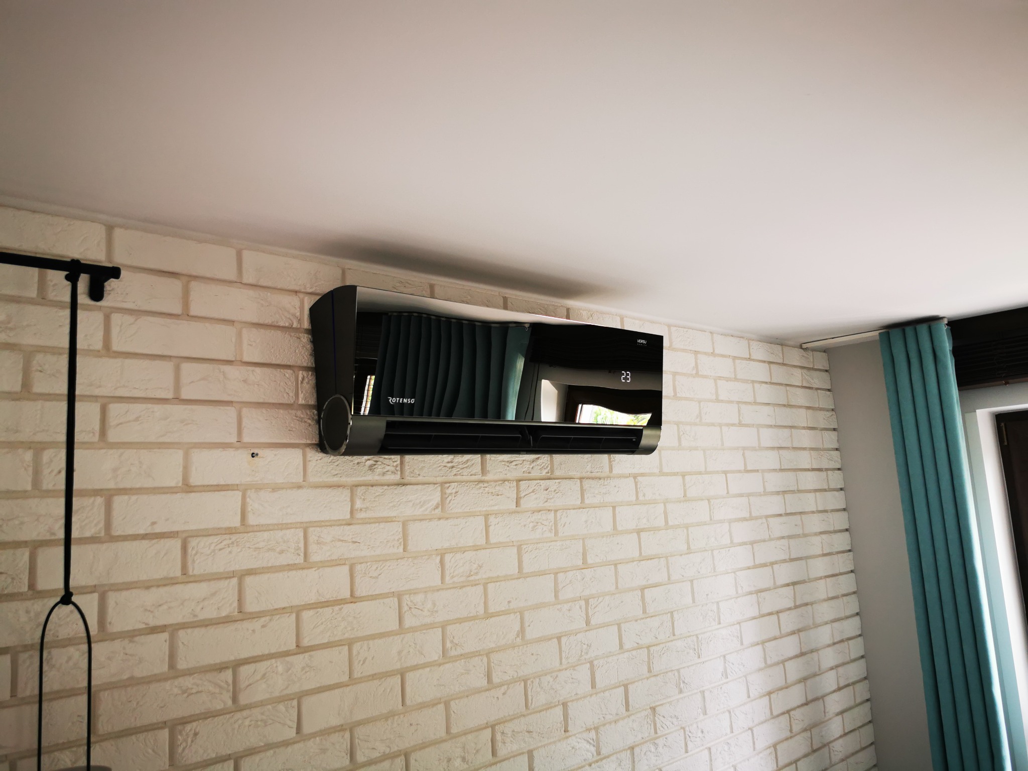 Czarny klimatyzator ścienny zamontowany na białej, ceglanej ścianie w pomieszczeniu z zasłonami w tle.