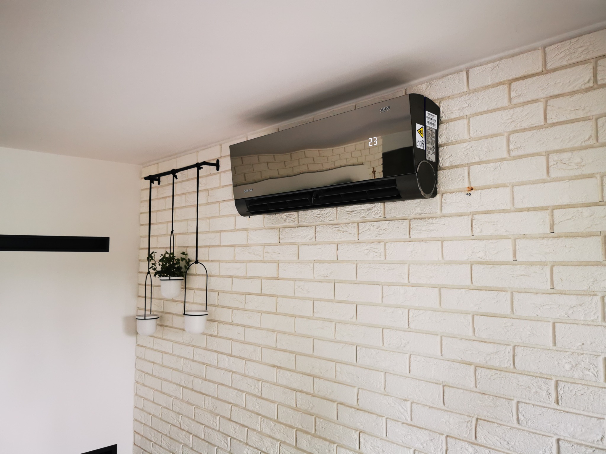 Czarny klimatyzator ścienny z wyświetlaczem temperatury (23 stopnie Celsjusza) na białej ścianie z cegły dekoracyjnej, obok wiszące doniczki z roślinami.