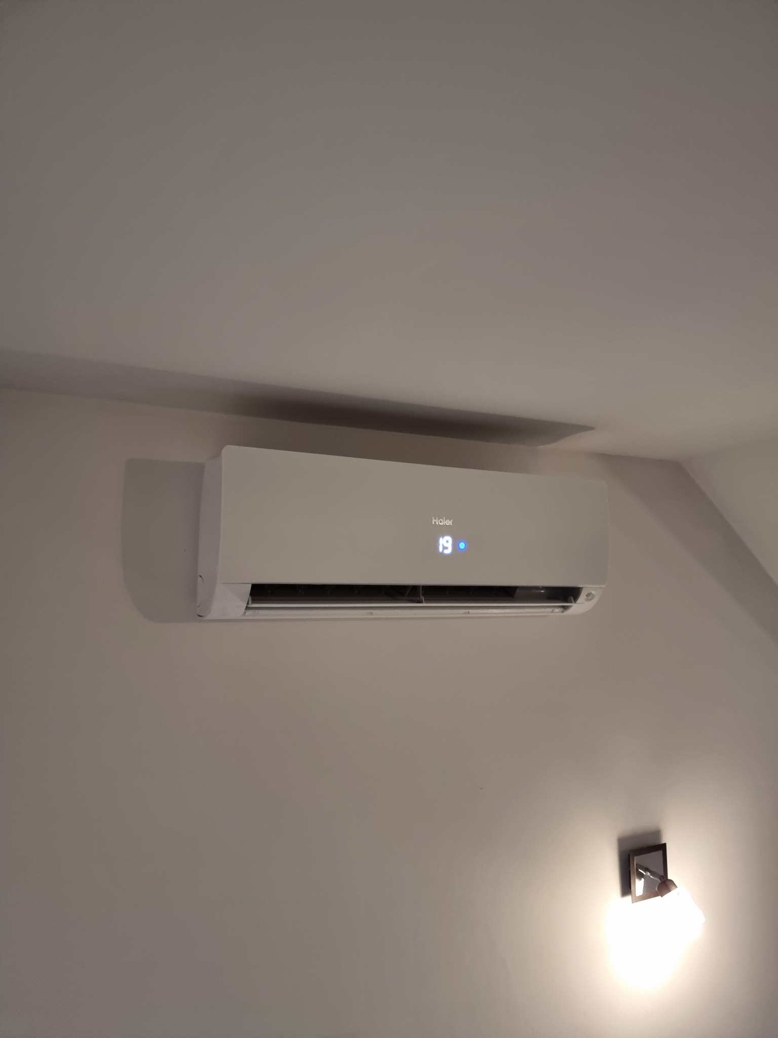 Biały klimatyzator Haier zamontowany na jasnej ścianie w pomieszczeniu, wyświetlający temperaturę 19 stopni Celsjusza, z widoczną lampką ścienną poniżej.