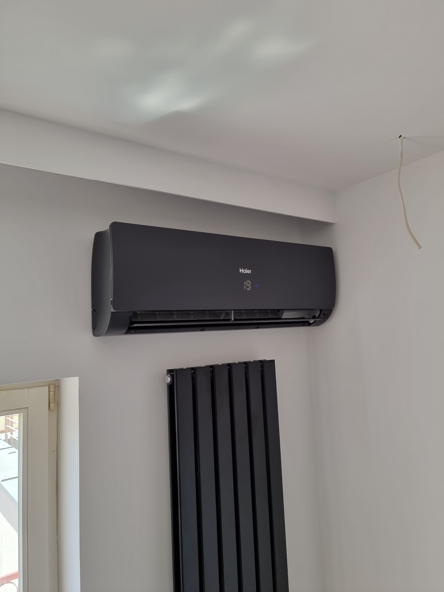 Czarny klimatyzator Haier z wyświetlaczem temperatury 19 stopni Celsjusza, zamontowany na białej ścianie w nowoczesnym wnętrzu, obok czarnego grzejnika panelowego i białego okna.