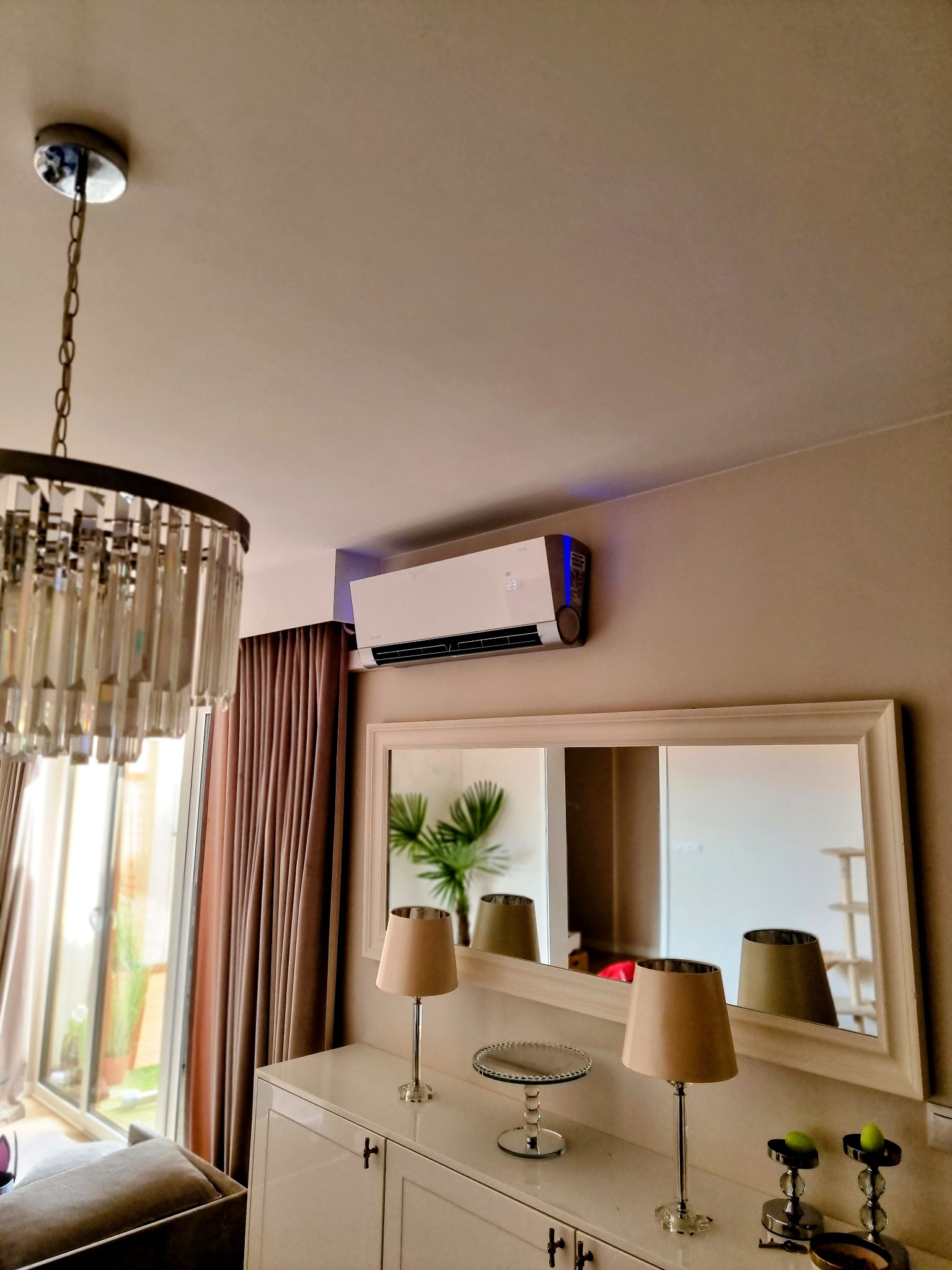 Nowoczesny klimatyzator ścienny z wyświetlaczem temperatury, zamontowany w eleganckim salonie z lustrem, lampami i żyrandolem.