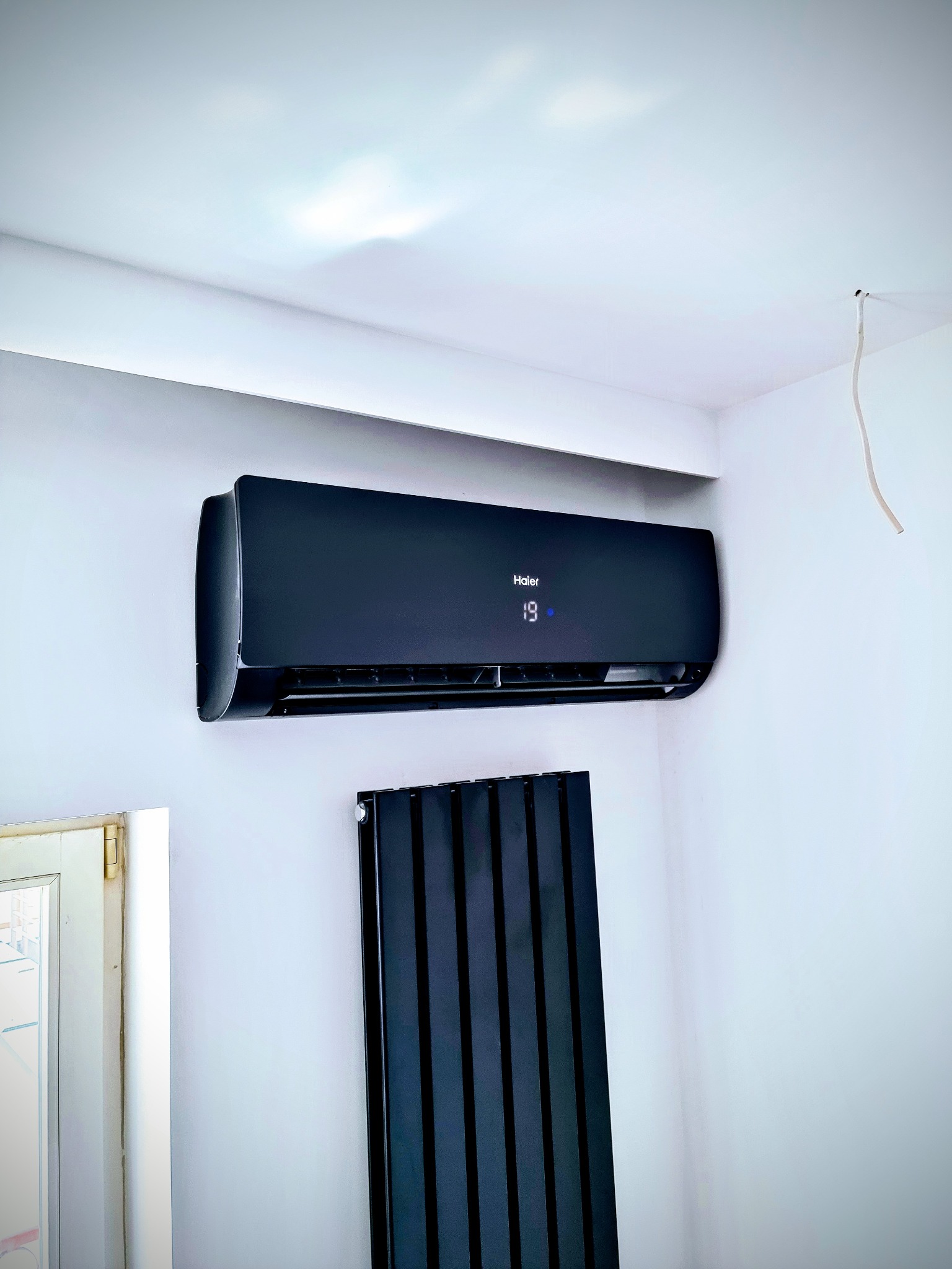 Czarny klimatyzator Haier zamontowany na białej ścianie w pomieszczeniu, obok grzejnika panelowego i fragmentu okna. Widoczny kabel wystający z sufitu.