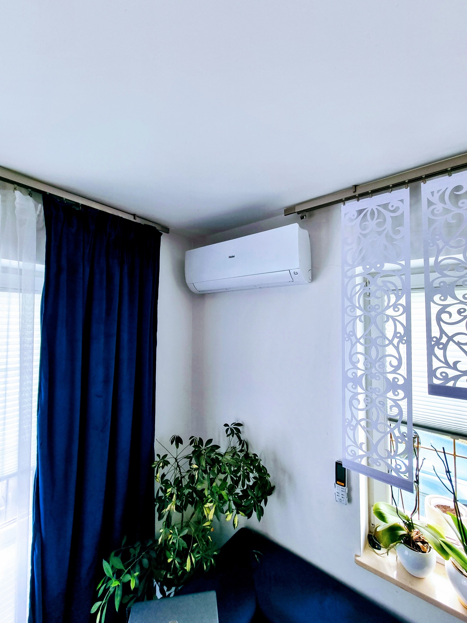 Biały klimatyzator marki Haier zamontowany na ścianie w pokoju z granatowymi zasłonami i dekoracyjnymi panelami okiennymi, widoczny pilot do klimatyzacji oraz rośliny doniczkowe.
