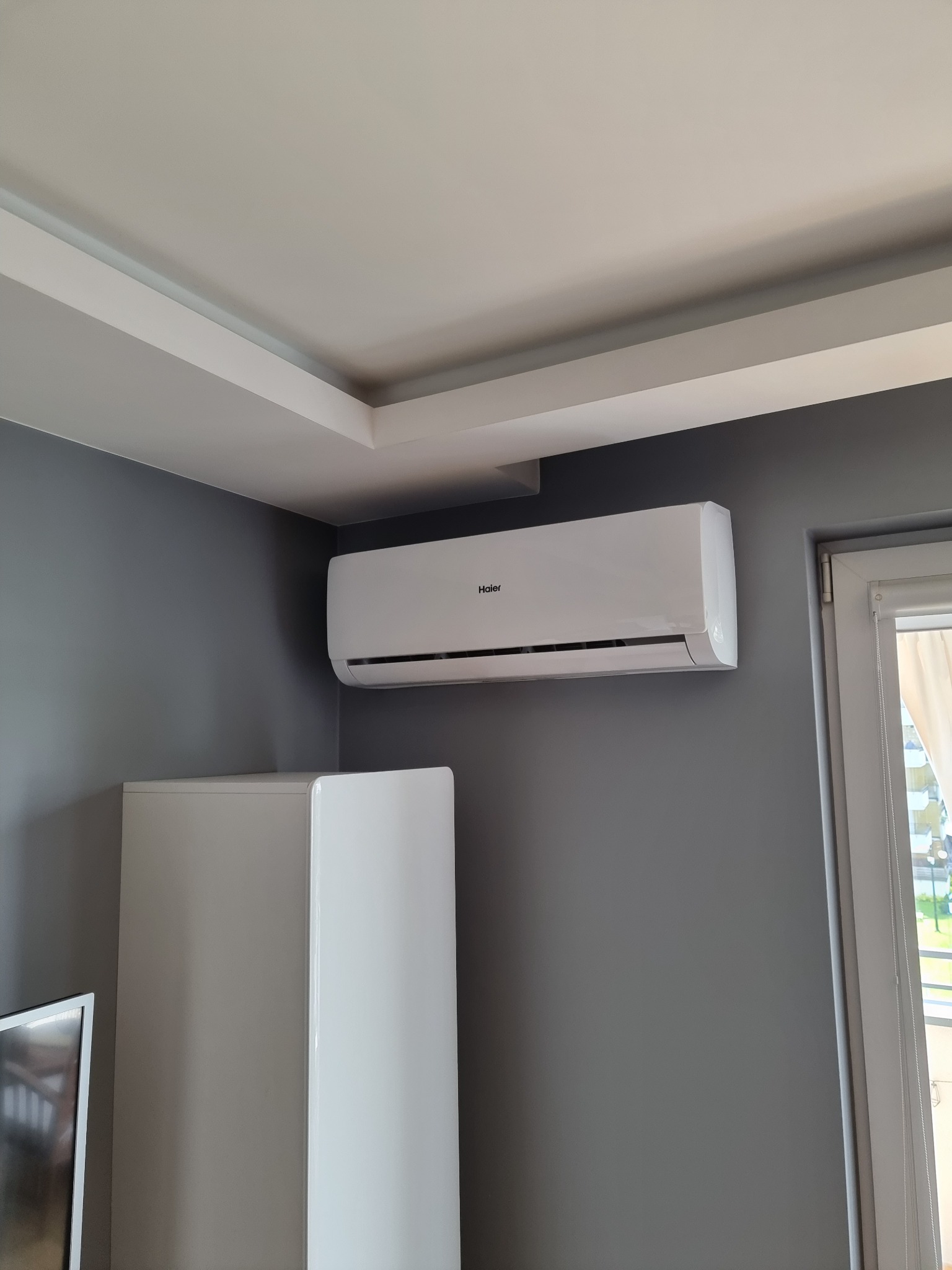 Biały klimatyzator Haier zamontowany na szarej ścianie w rogu pokoju, obok okna z zasłoną i wysokiej białej szafki.