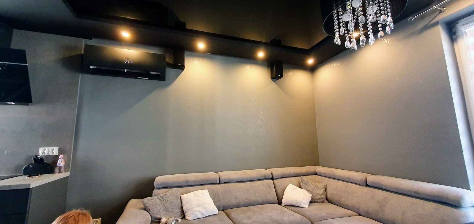 Czarny klimatyzator ścienny Haier z wyświetlaczem temperatury 23 stopnie Celsjusza, zamontowany w nowoczesnym salonie z szarą ścianą, oświetleniem punktowym i kryształowym żyrandolem nad narożną sofą.