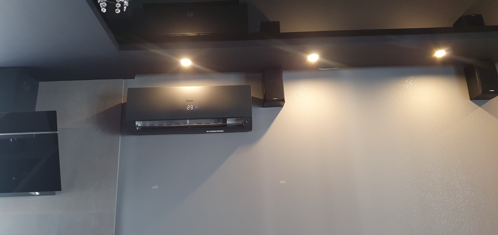 Czarny klimatyzator Haier zamontowany na szarej ścianie w nowoczesnym wnętrzu, widoczny wyświetlacz temperatury 23 stopnie Celsjusza, oświetlenie punktowe na suficie.