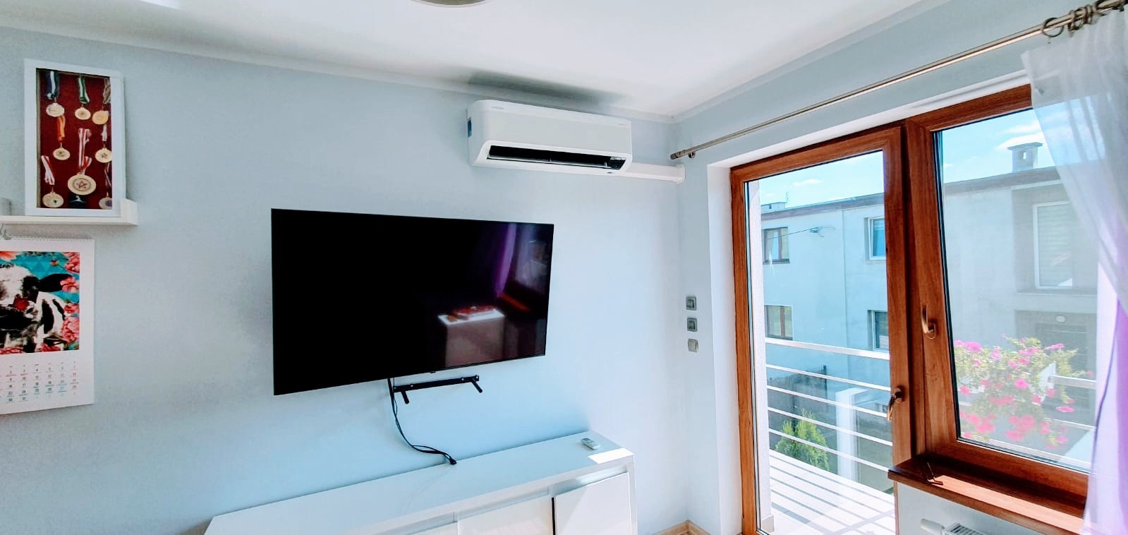 Nowoczesny klimatyzator ścienny w jasnym pokoju z telewizorem, widok na balkon i budynek na zewnątrz.
