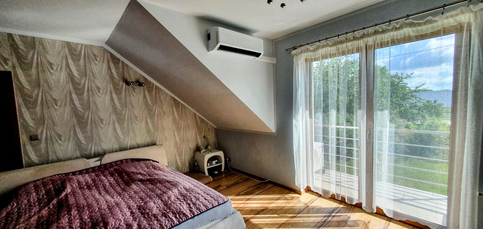 Biała jednostka klimatyzacyjna zamontowana na skośnej ścianie w sypialni na poddaszu z wzorzystą tapetą i dużym oknem balkonowym z firankami.