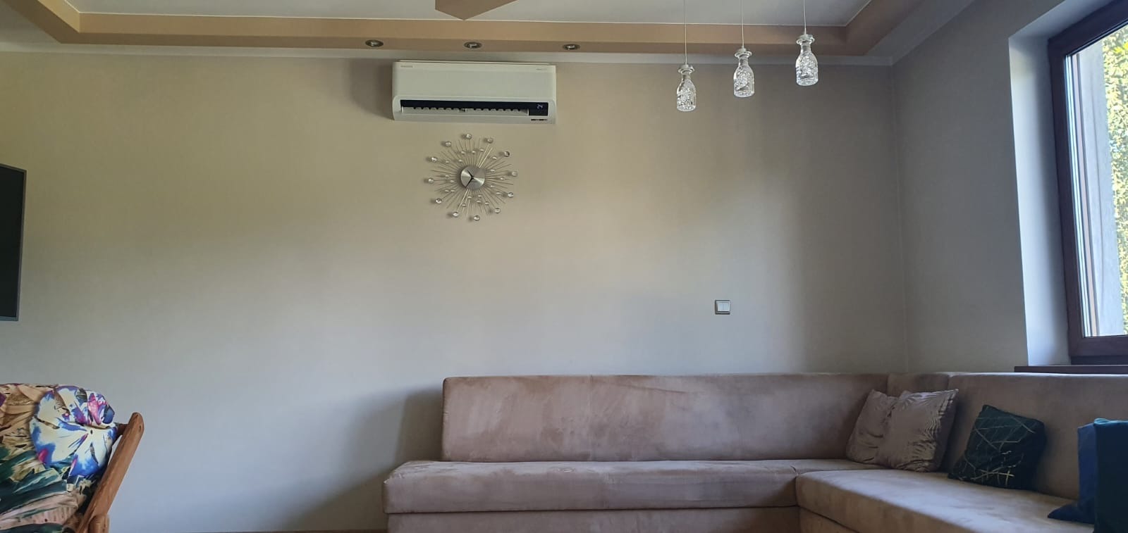Biały klimatyzator ścienny zamontowany w salonie nad beżową sofą narożną, ozdobiony dekoracyjnym zegarem z kryształkami i trzema szklanymi lampami wiszącymi.