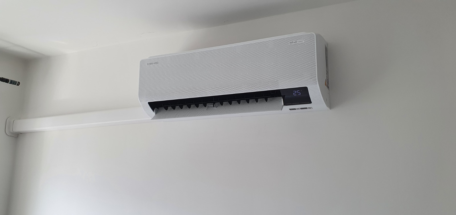 Biały klimatyzator Samsung WindFree zamontowany na białej ścianie, widoczna rura instalacyjna i wyświetlacz pokazujący temperaturę 25 stopni.