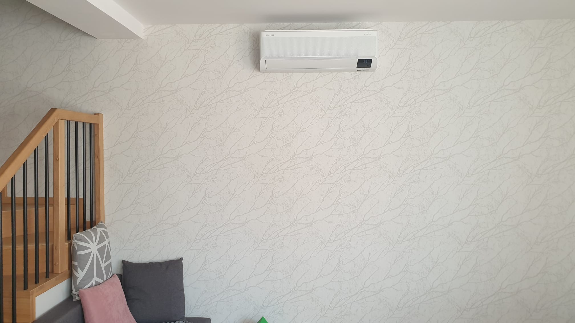 Biały klimatyzator Samsung zamontowany na ścianie z delikatnym wzorem gałązek w salonie z drewnianymi schodami i szarą sofą z poduszkami.