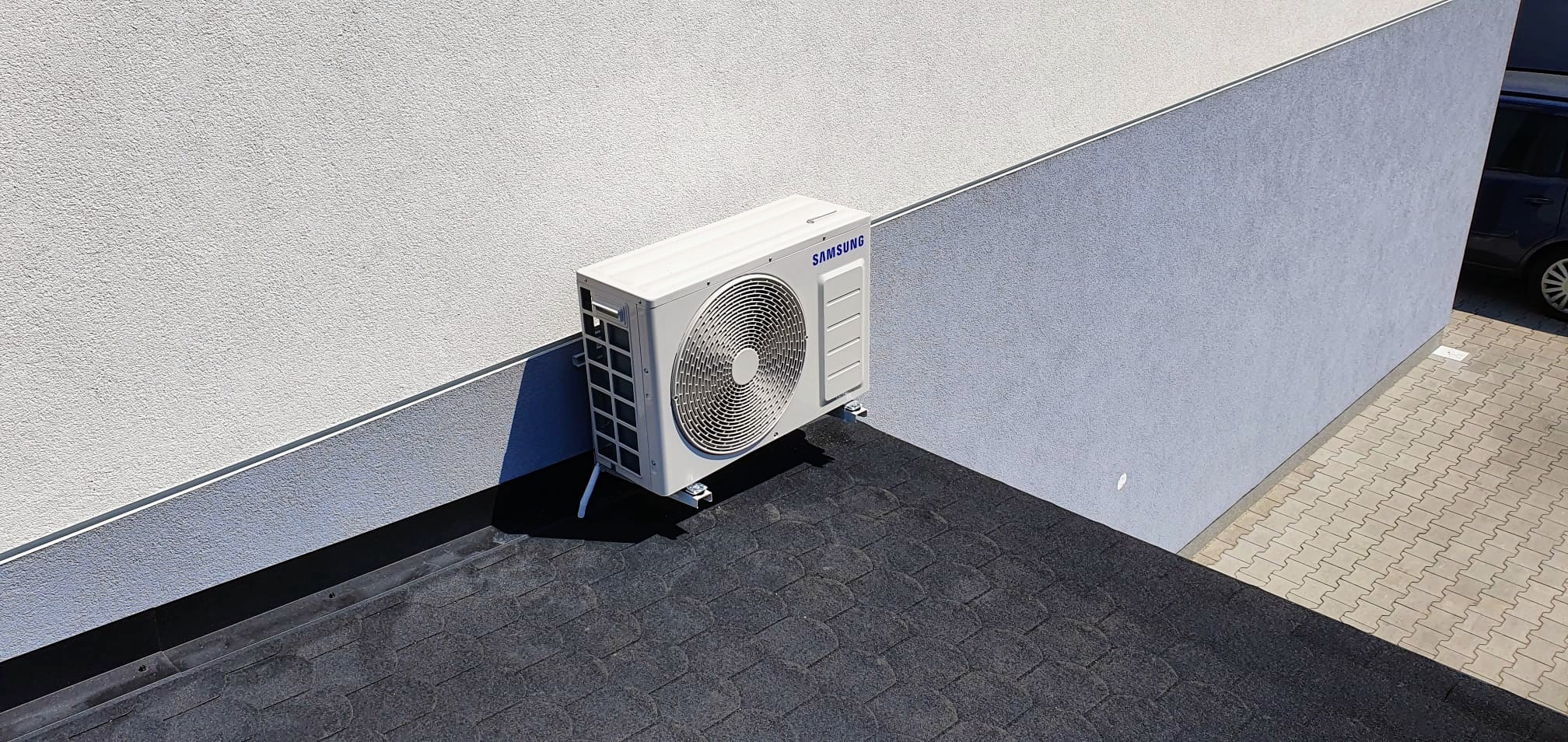 Biała jednostka zewnętrzna klimatyzacji Samsung zamontowana na dachu pokrytym czarnym gontem bitumicznym, przylegająca do ściany budynku z dwoma odcieniami szarości.
