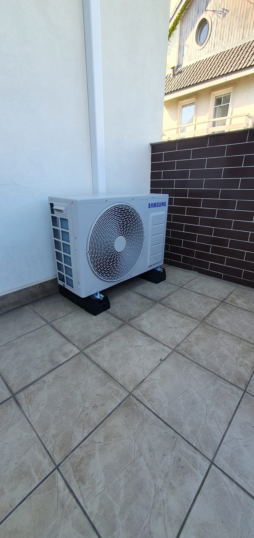 Zewnętrzna jednostka klimatyzacji Samsung zainstalowana na balkonie w Kowali Stępocinie, umieszczona na czarnych, gumowych podkładkach antywibracyjnych, z widoczną ścianą z brązowej cegły w tle.
