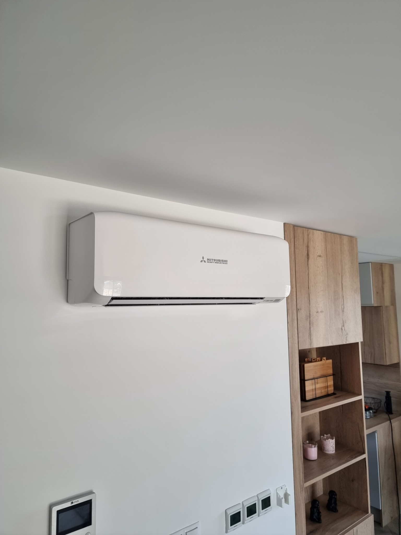 Biały klimatyzator Mitsubishi zamontowany na ścianie w nowoczesnym wnętrzu, obok drewnianych półek z dekoracjami.