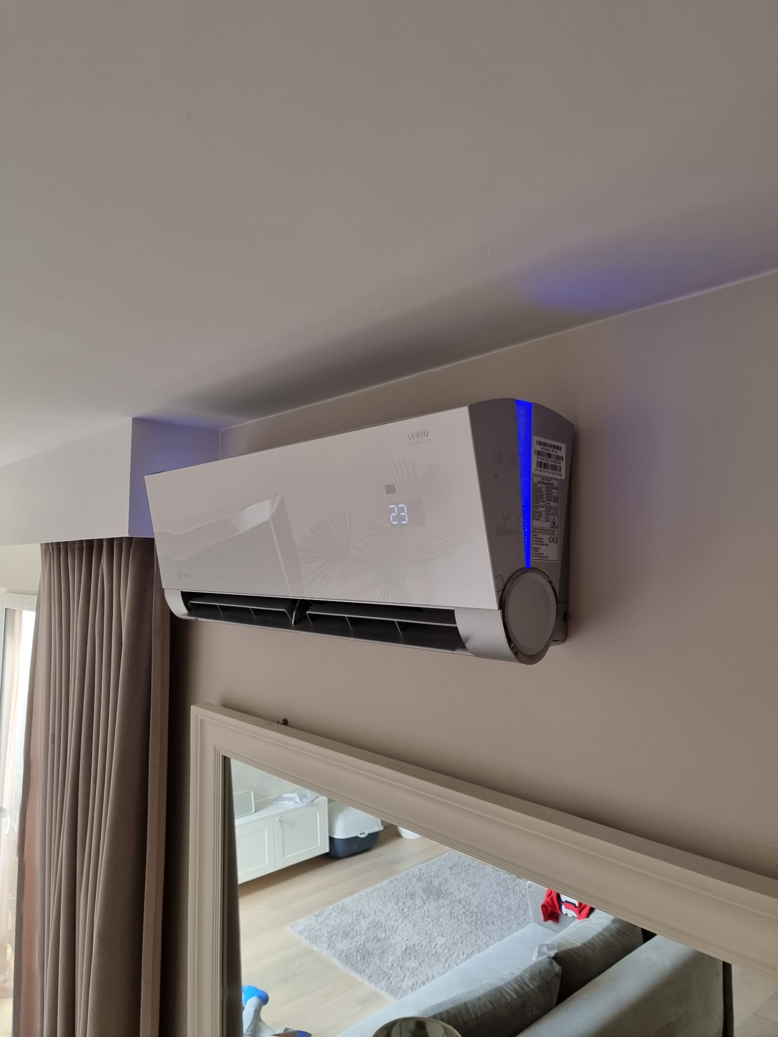 Biały klimatyzator marki Versu z wyświetlaczem temperatury 23 stopnie Celsjusza, zamontowany na ścianie w pokoju, widoczny w odbiciu lustra fragment sofy i okna z zasłonami.
