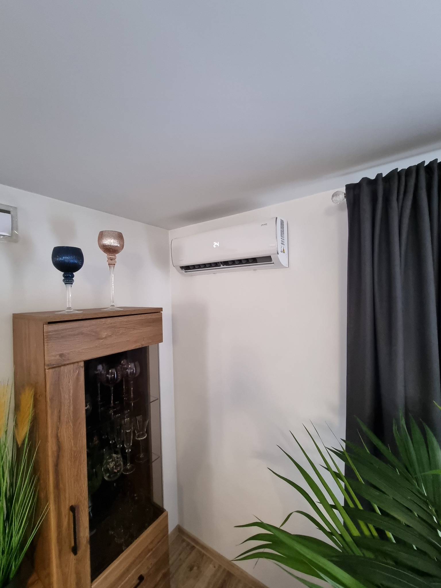 Biały klimatyzator ścienny włączony, wyświetlający temperaturę 24 stopnie Celsjusza, zamontowany w salonie z brązową szafką, dekoracyjnymi kielichami i zasłonami.