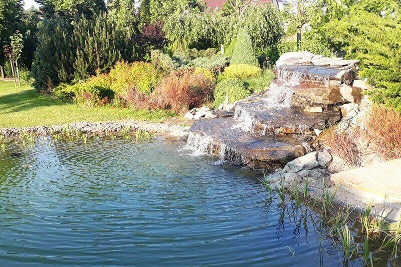 Oczko wodne z kaskadą wodną wykonaną z kamieni, otoczone zielenią i roślinnością, z widocznym fragmentem drewnianego podestu.