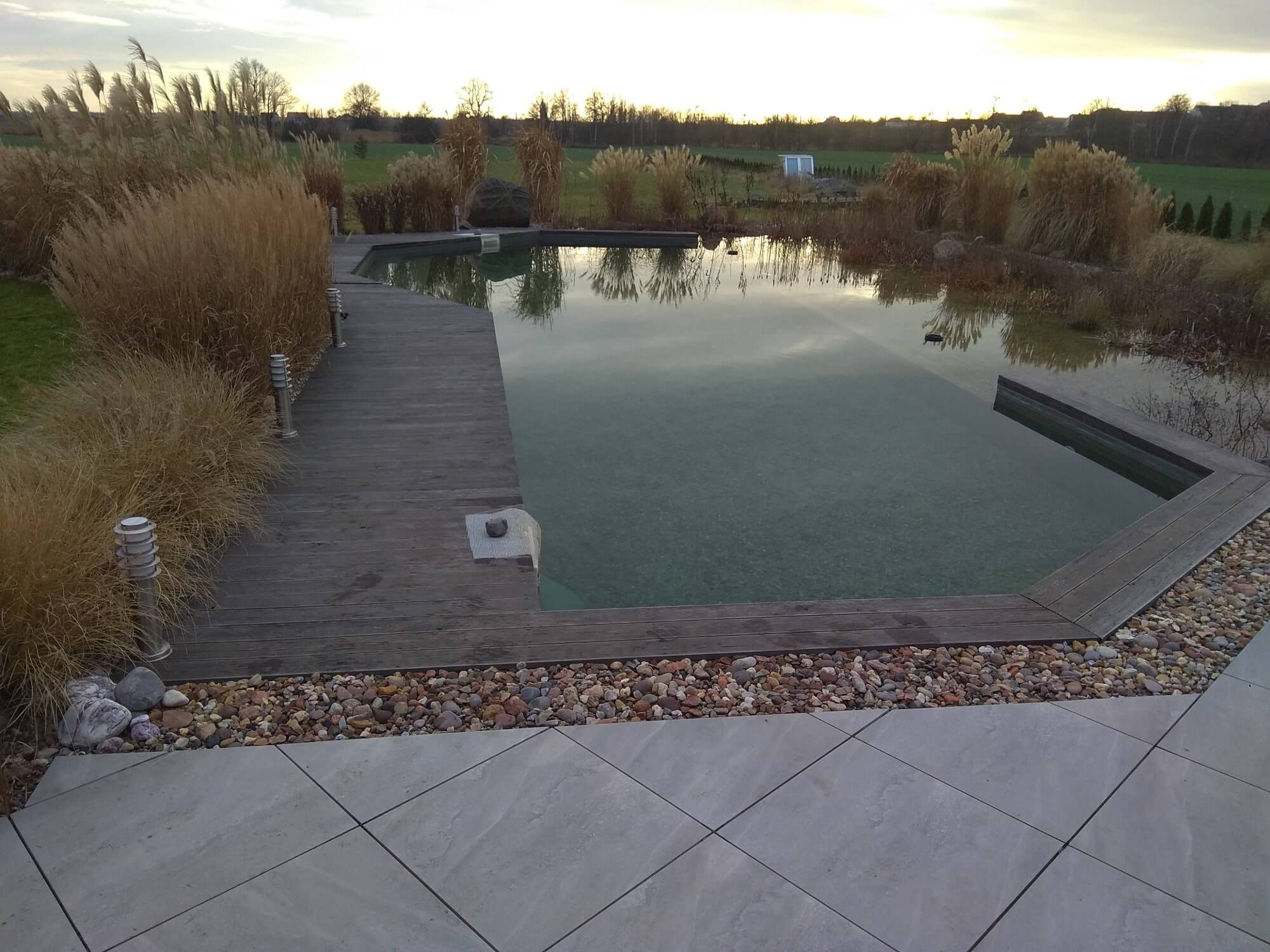 Naturalny zbiornik wodny we Wrocławiu, z drewnianym pomostem i kamienną obudową, otoczony ozdobnymi trawami, tworzący relaksującą strefę wypoczynku w ogrodzie.