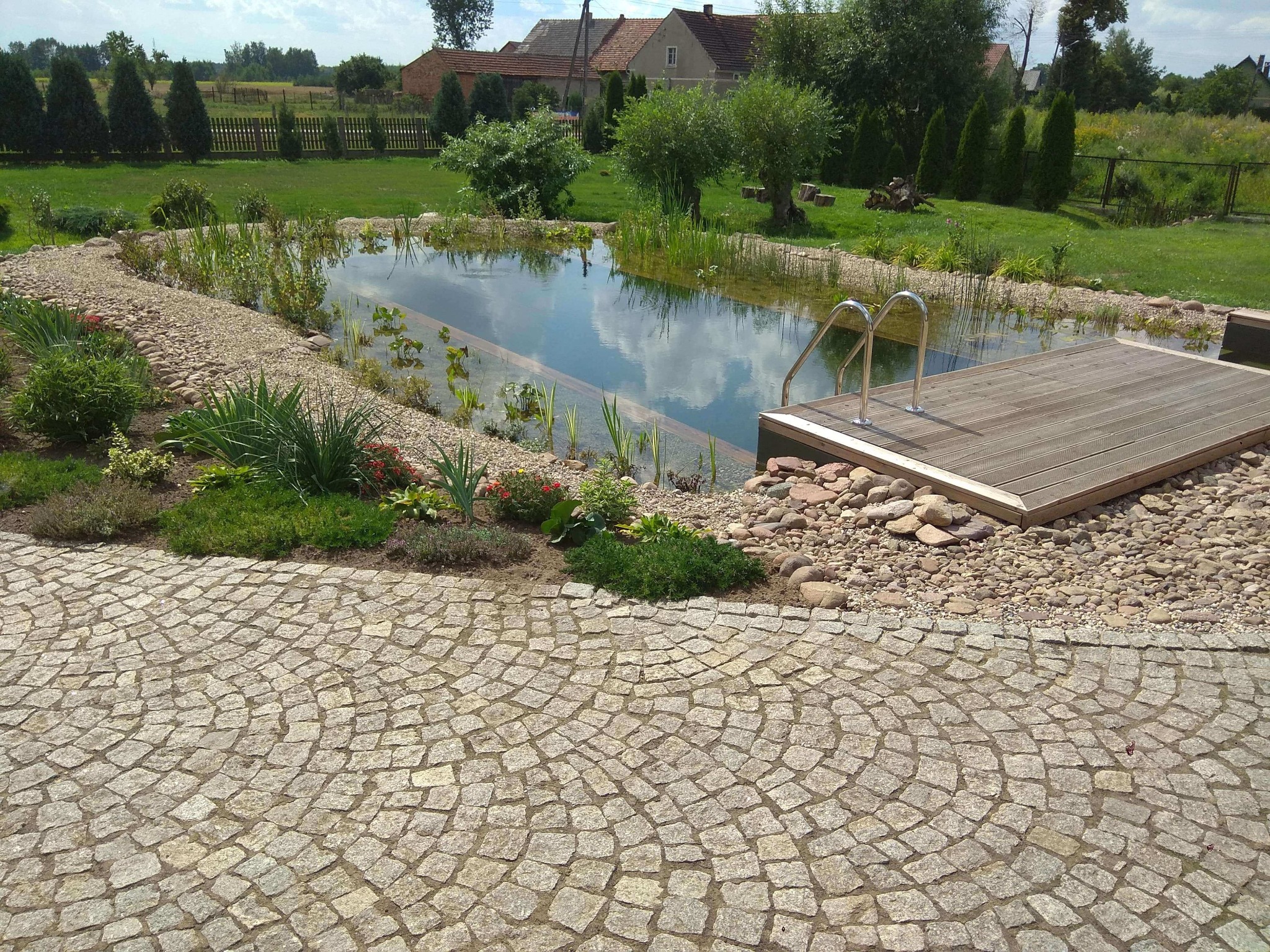 Naturalny zbiornik wodny z drewnianym pomostem i metalową drabinką, otoczony roślinnością i ścieżką z kostki granitowej ułożonej w wachlarz, na tle wiejskich zabudowań.