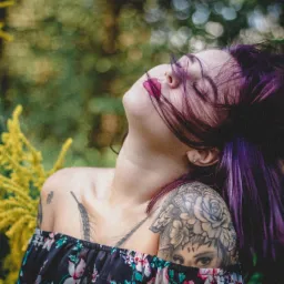 Portret kobiety z fioletowymi włosami i tatuażami na ramionach, odchylonej głowy do tyłu na tle rozmytej zieleni i żółtych kwiatów.