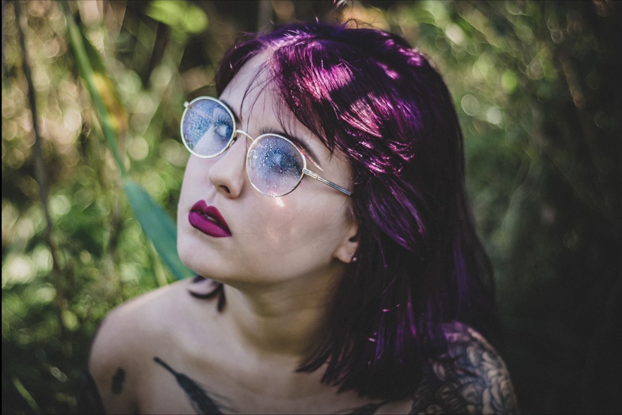 Portret kobiety z fioletowymi włosami i okrągłymi okularami, spoglądającej w górę na tle zieleni