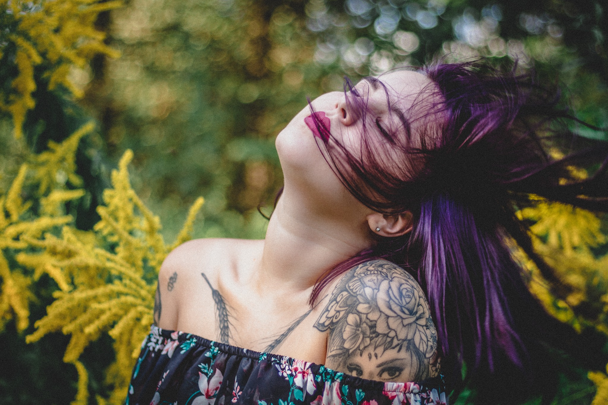 Portret kobiety z fioletowymi włosami i tatuażami na ramionach, odchylonej głowy do tyłu na tle rozmytej zieleni i żółtych kwiatów.