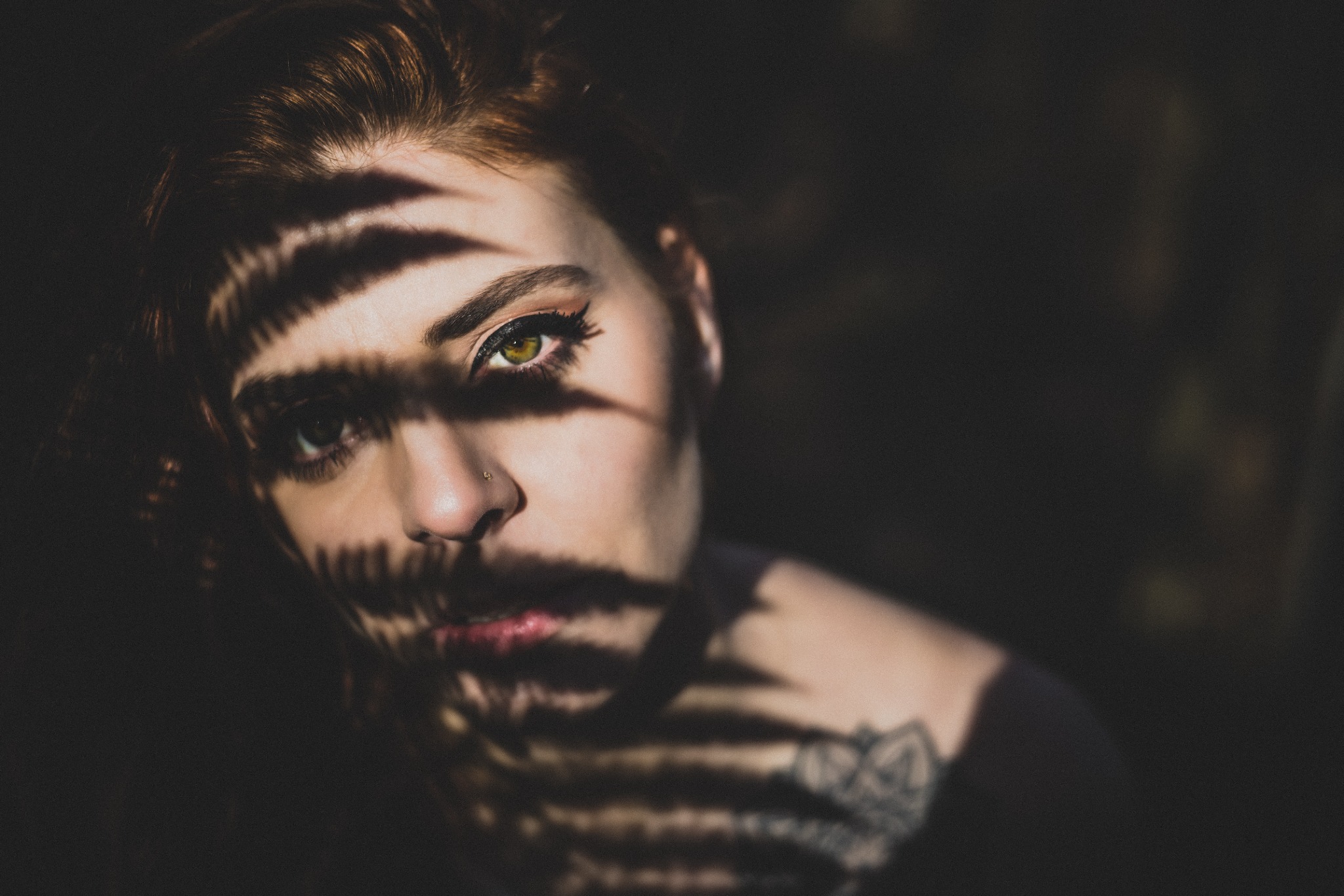 Portret kobiety z rudymi włosami, z zielonymi oczami i piercingiem w nosie, oświetlonej cieniem liści paproci, z widocznym tatuażem na ramieniu.