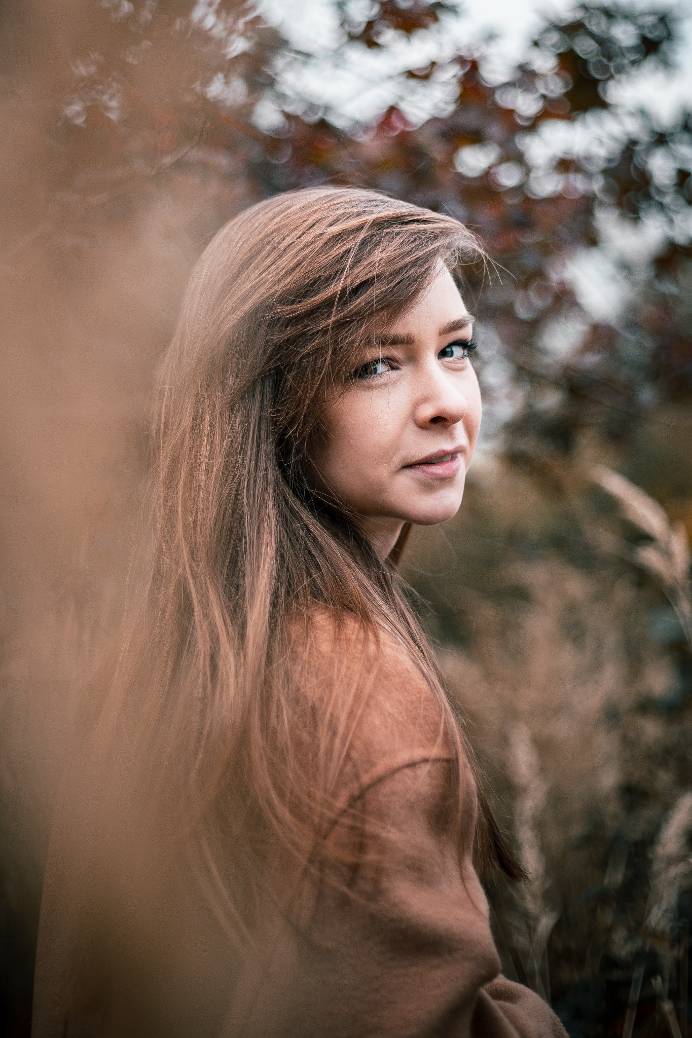 Portret młodej kobiety z długimi, brązowymi włosami, spoglądającej przez ramię w zamyślonym wyrazem twarzy, na tle rozmytej roślinności w jesiennych barwach.