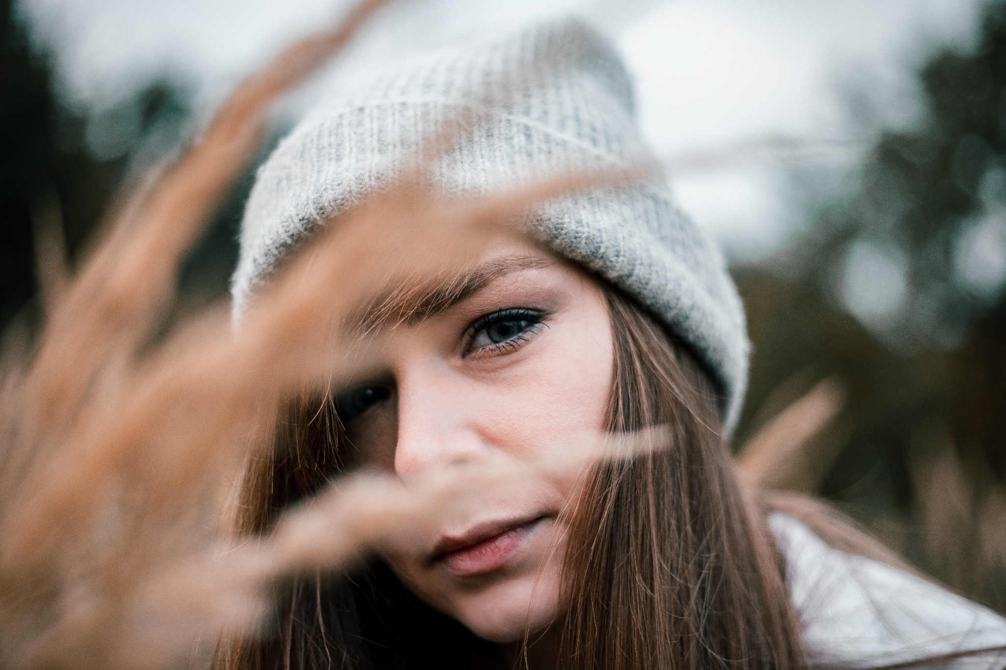 Portret młodej kobiety w szarej czapce, spoglądającej przez brązowe trawy, z delikatnym uśmiechem i wyrazistym makijażem oczu.