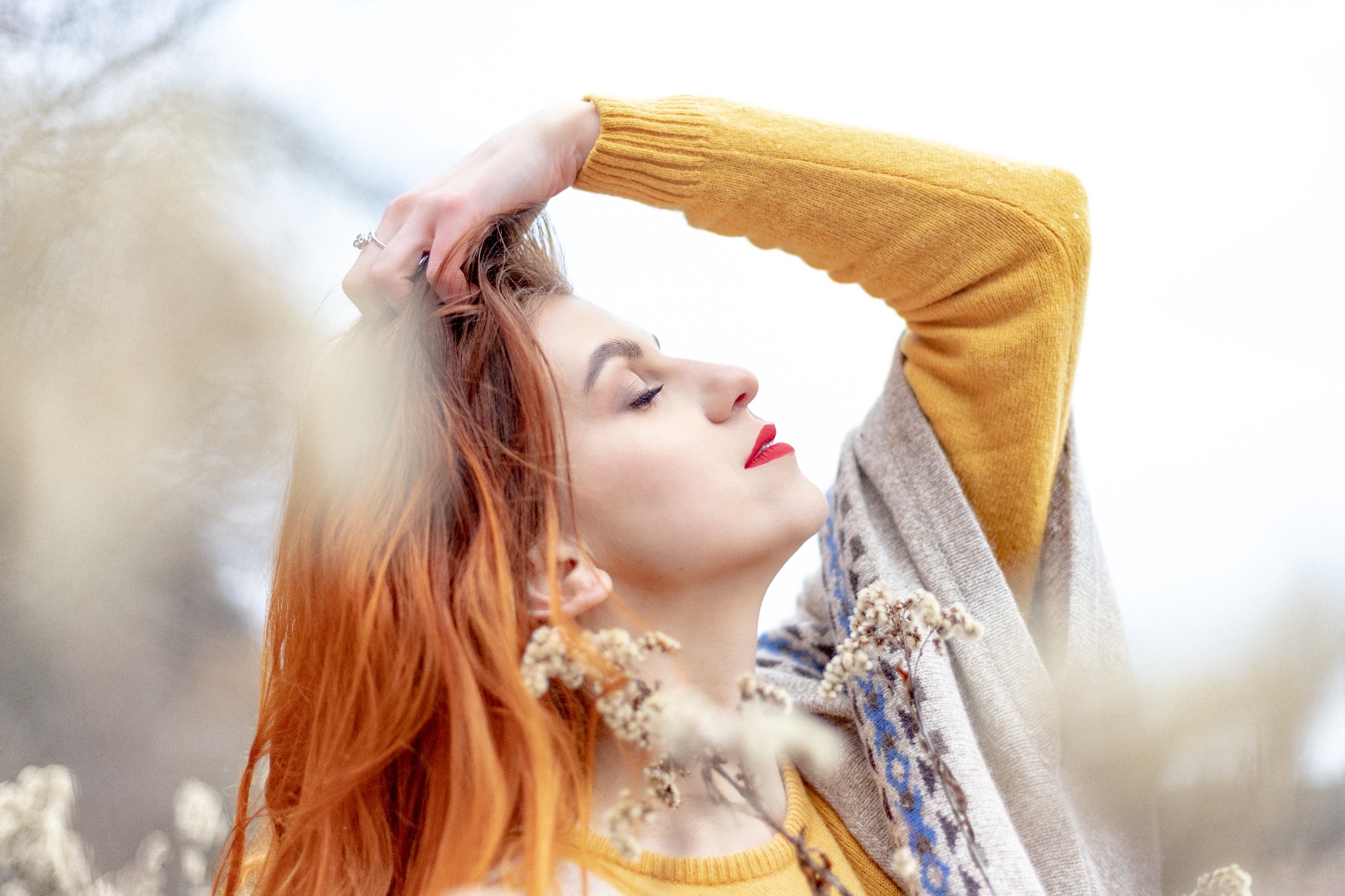 Portret kobiety z rudymi włosami w żółtym swetrze i szalu, z zamkniętymi oczami i czerwonymi ustami, pozującej na tle rozmytej roślinności.
