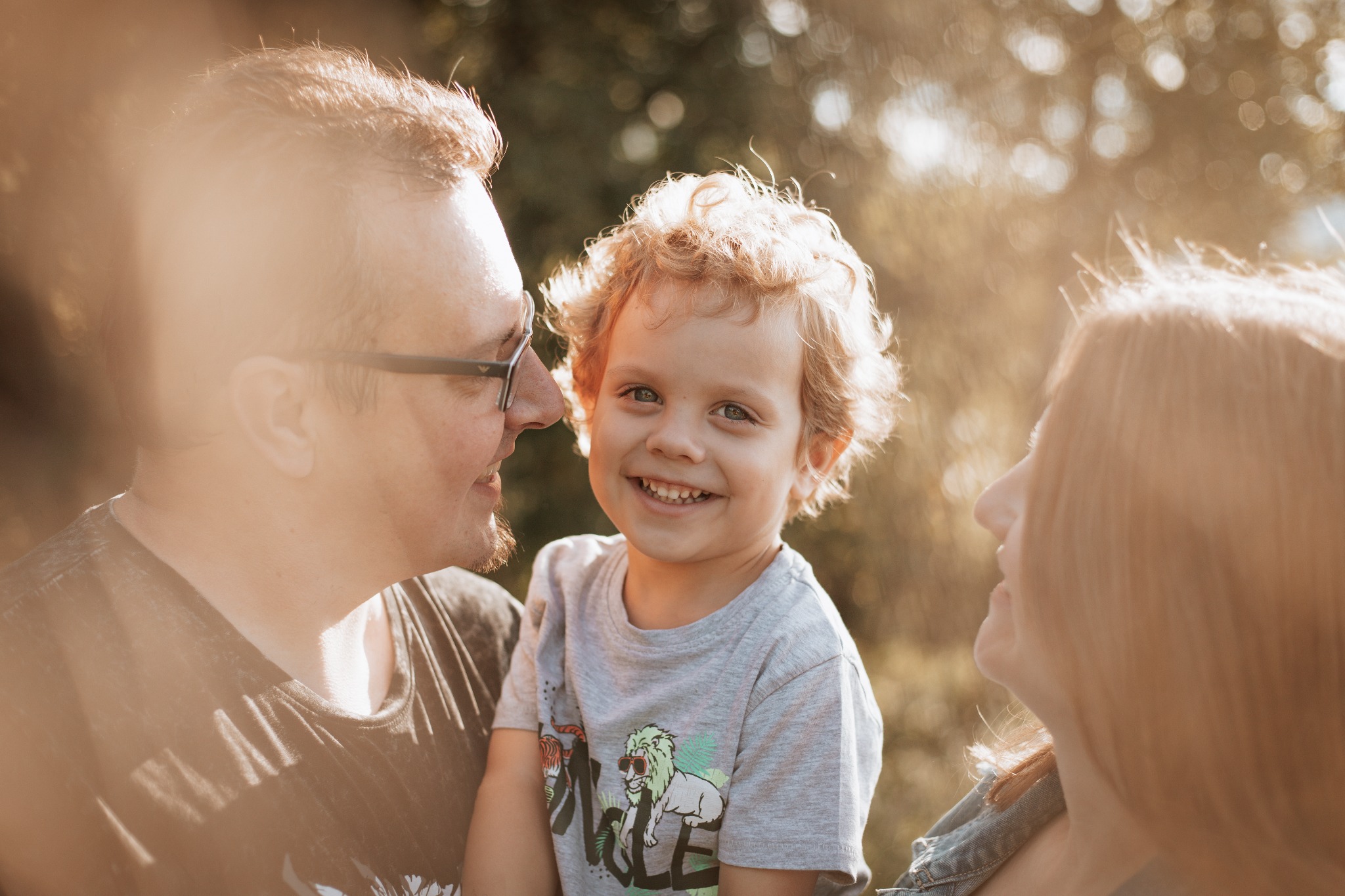 Uśmiechnięty chłopiec z kręconymi włosami na tle rodziców, ciepłe światło słoneczne, naturalna sceneria.