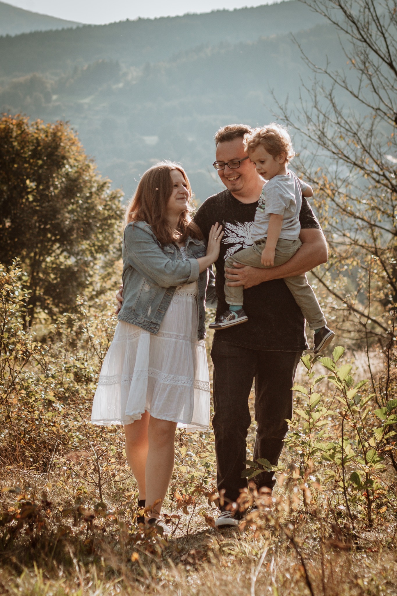 Uśmiechnięta rodzina: mama w białej sukience i dżinsowej kurtce, tata w okularach trzymający na rękach synka w plenerze z zalesionym wzgórzem w tle.