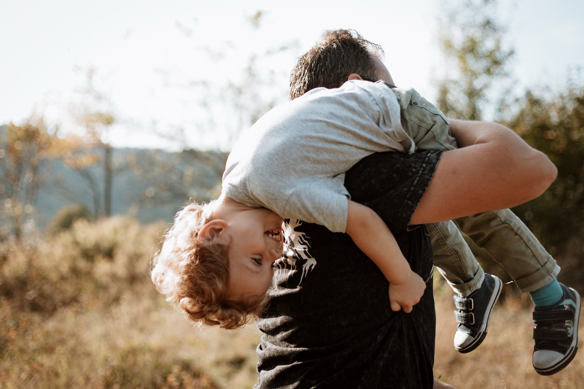 Ojciec trzymający syna do góry nogami na tle łąki w słoneczny dzień, dziecko się śmieje.
