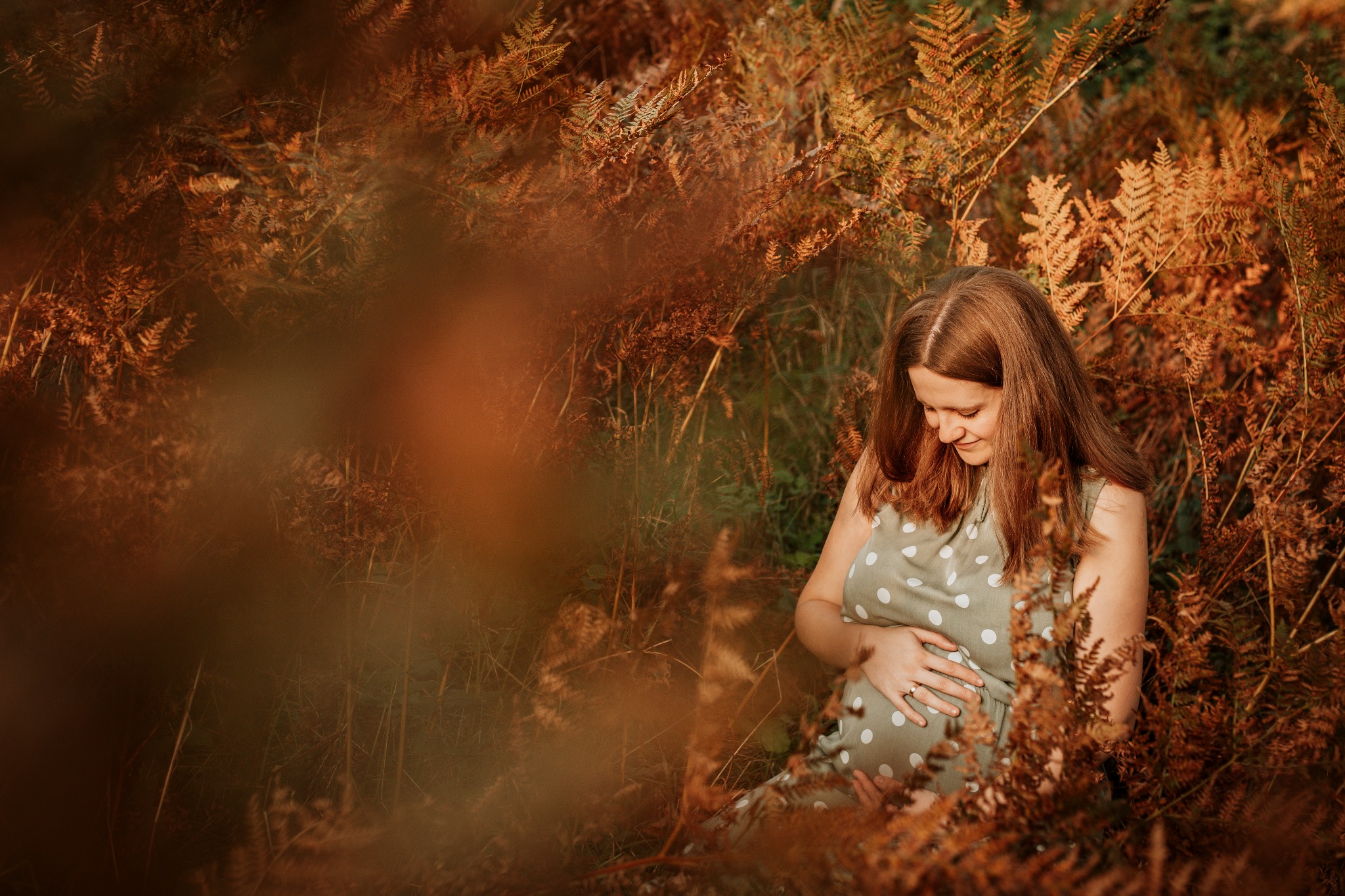 Kobieta w ciąży w sukience w groszki, otoczona brązowymi paprociami, delikatne światło podkreśla naturalność sceny.