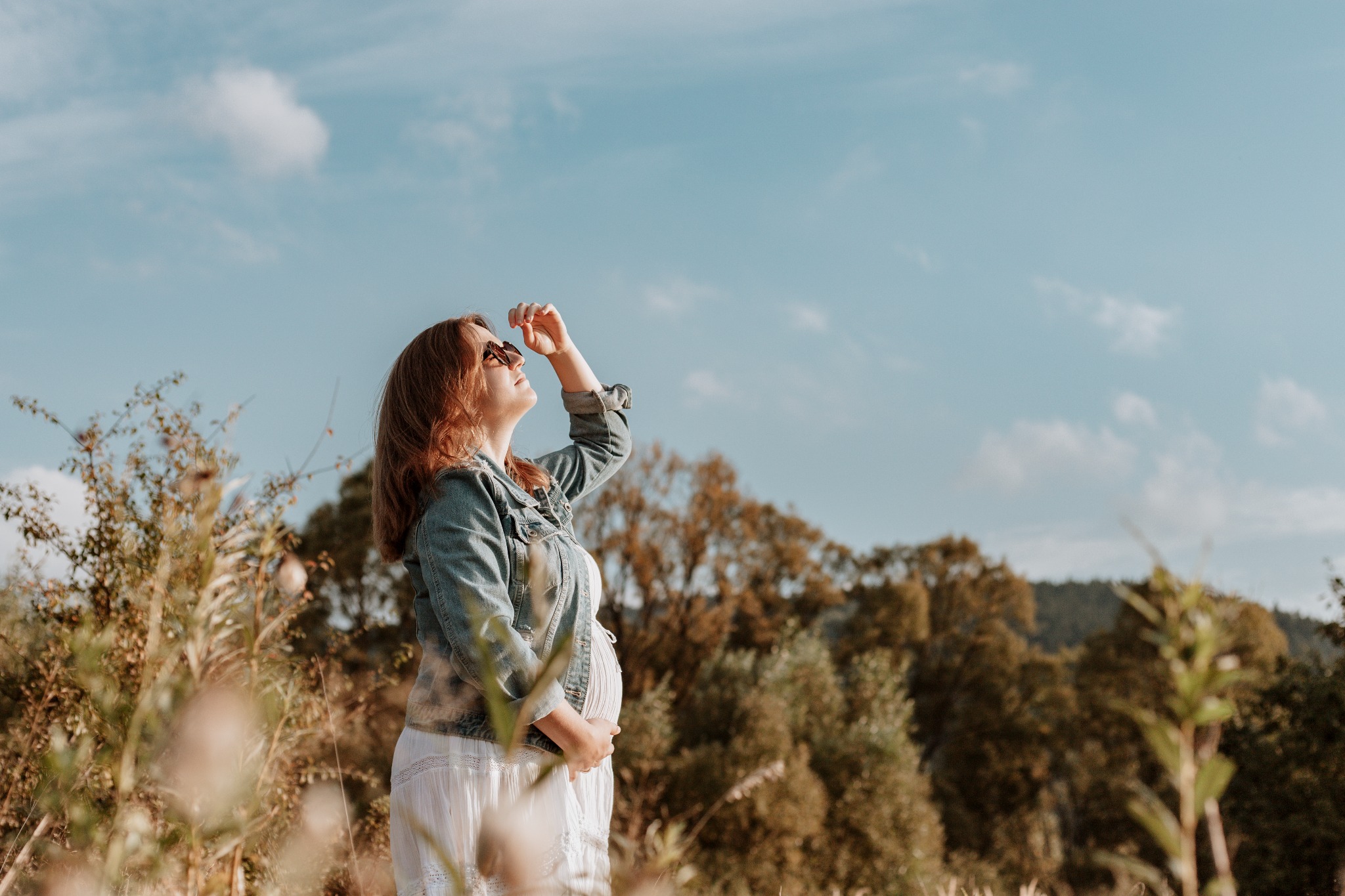 Kobieta w ciąży w białej sukience i jeansowej kurtce, stojąca na łące, patrzy w górę na niebo, trzymając dłoń nad okularami przeciwsłonecznymi.