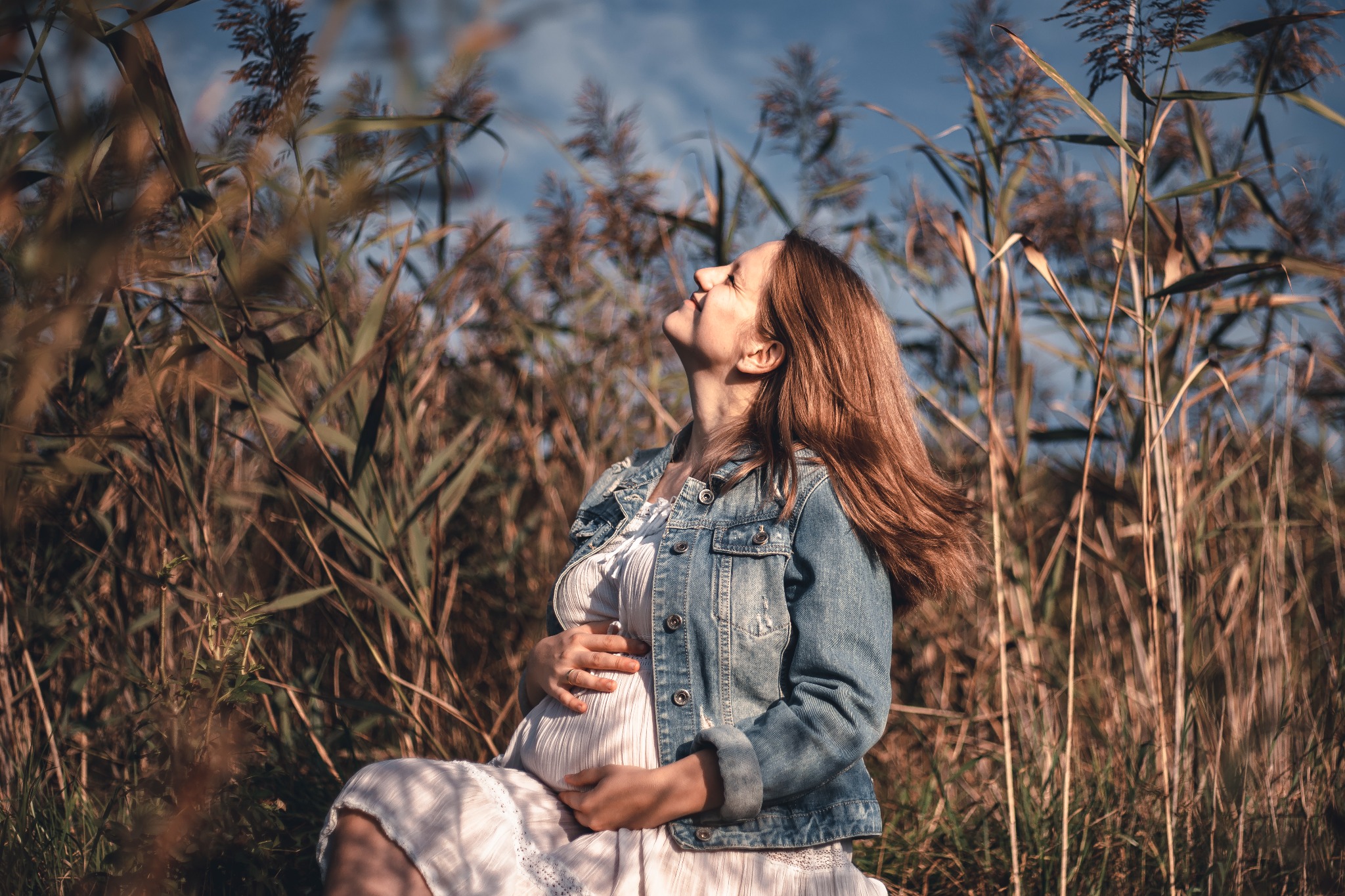 Kobieta w ciąży w denimowej kurtce i białej sukience siedzi w trawie, spoglądając w górę na błękitne niebo, otoczona wysokimi trawami i trzcinami.