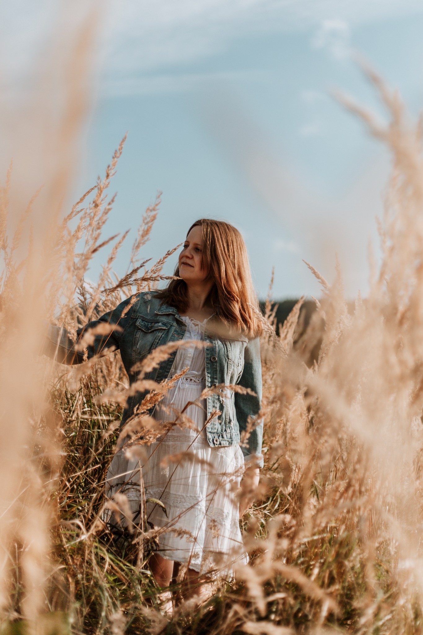 Kobieta w białej sukience i jeansowej kurtce stoi na polu wysokiej trawy, spoglądając w dal na tle błękitnego nieba.