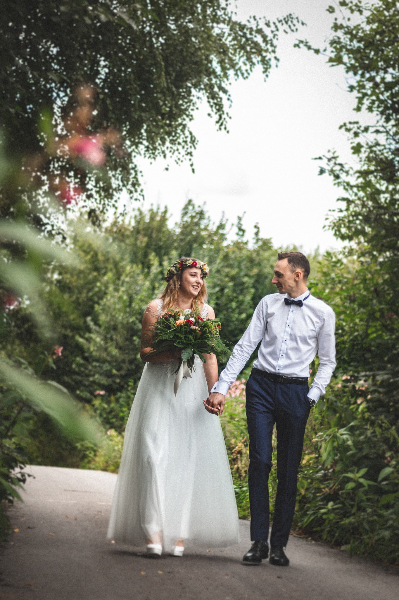 Para młoda spaceruje ścieżką w lesie, panna młoda w białej sukni z bukietem i wiankiem, pan młody w granatowych spodniach i białej koszuli z muszką, trzymają się za ręce i patrzą na siebie.