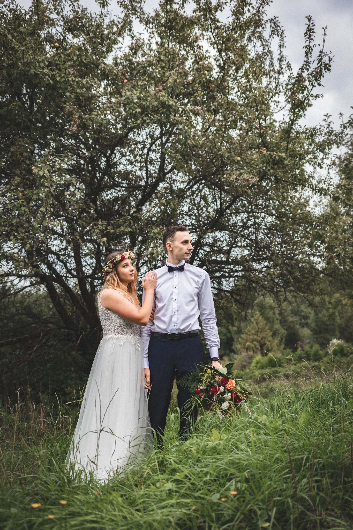 Para młoda w plenerze: panna młoda w wianku z kwiatów opiera dłoń na ramieniu pana młodego z bukietem, na tle drzewa i wysokiej trawy.