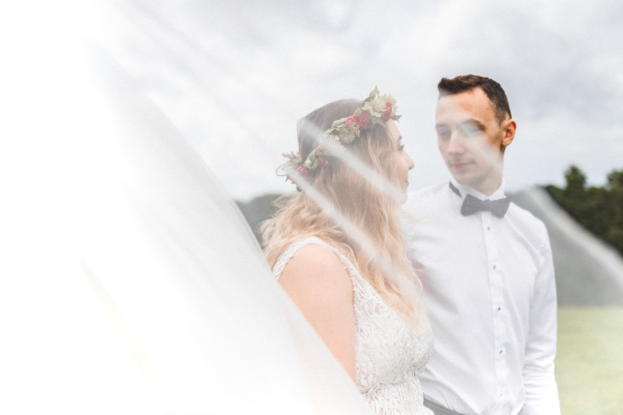 Para młoda w plenerze, panna młoda w koronie z kwiatów i sukni z koronki, pan młody w muszce, oboje patrzą na siebie przez welon.