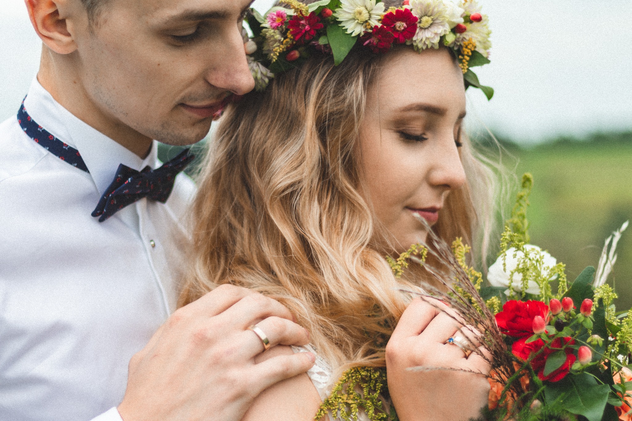 Młoda para w plenerze: panna młoda z wiankiem z polnych kwiatów i bukietem, pan młody w białej koszuli z muszką, delikatny uścisk, złote obrączki.