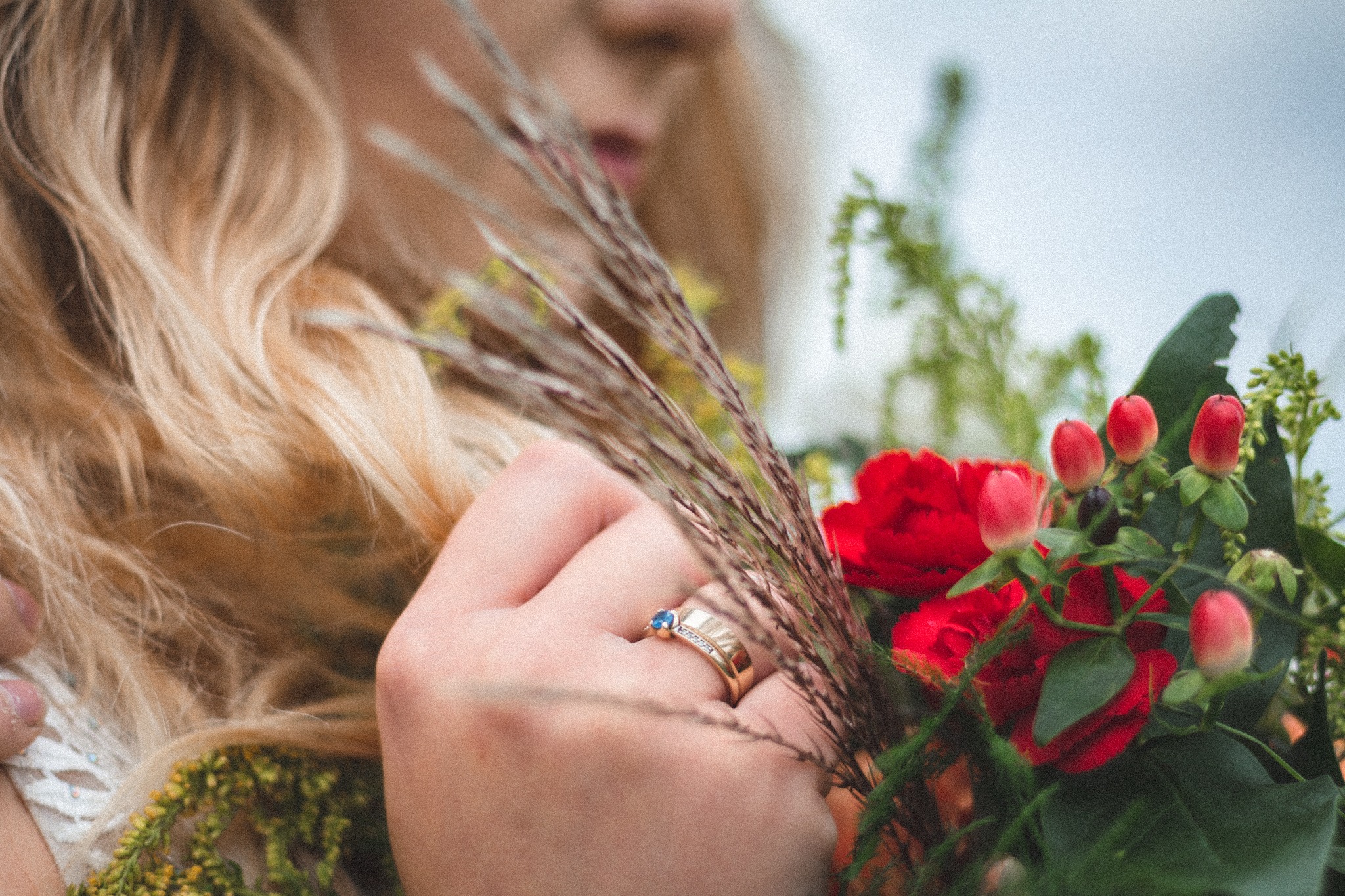 Dłoń kobiety z pierścionkiem z niebieskim oczkiem trzymająca bukiet czerwonych róż i traw, widoczny fragment blond włosów i białej sukienki.