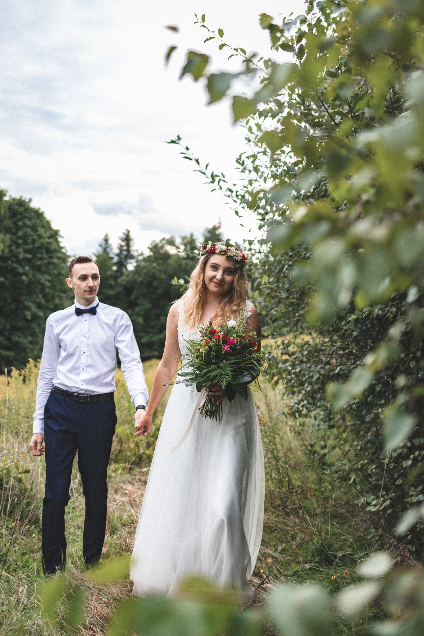 Para młoda trzymająca się za ręce spaceruje pośród zieleni, panna młoda w białej sukni z bukietem i wiankiem na głowie, pan młody w białej koszuli z muchą.