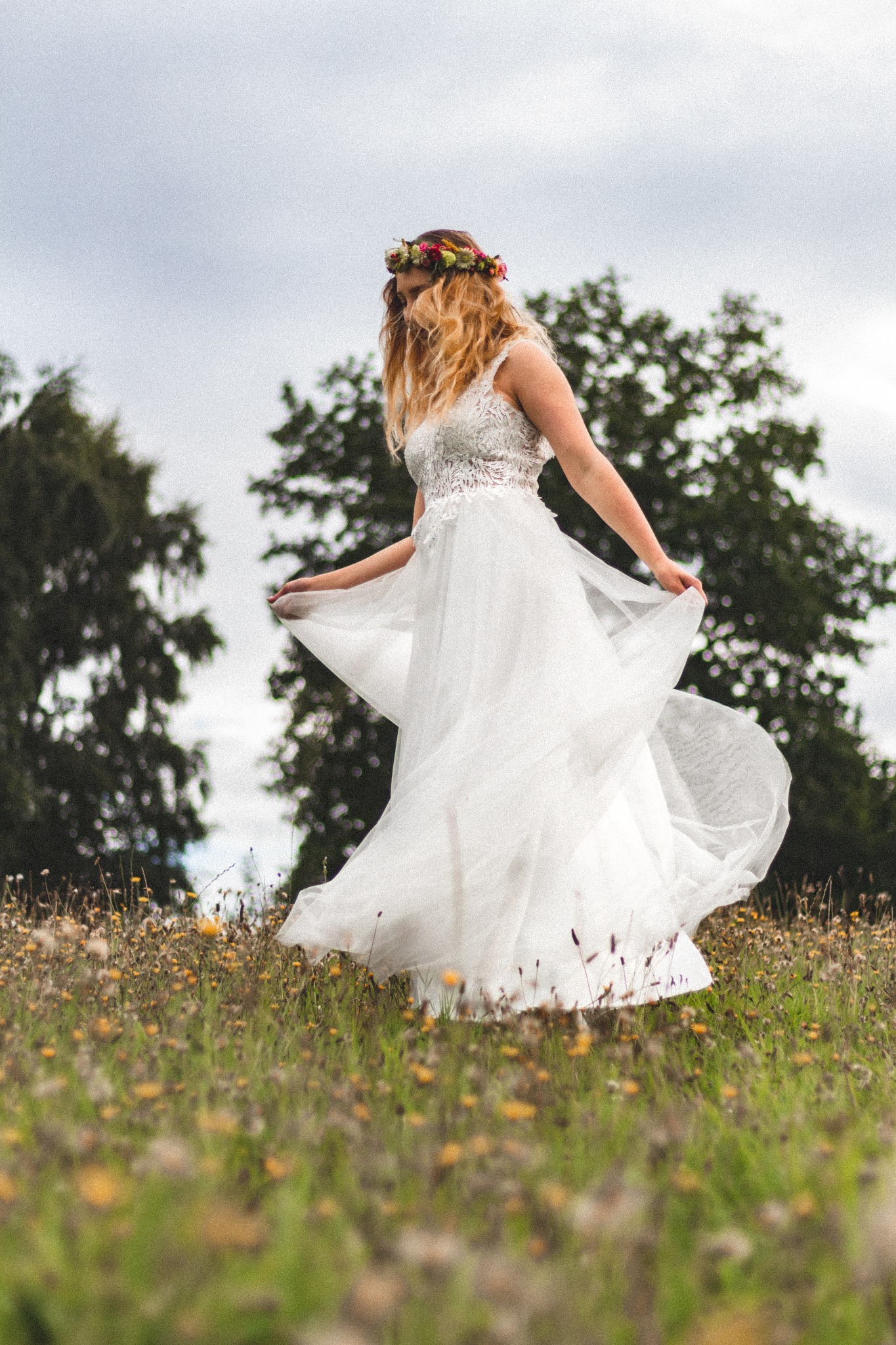 Kobieta w białej sukni z koronkową górą i kwiatowym wiankiem na głowie, tańcząca na łące pełnej żółtych kwiatów, z drzewami w tle pod pochmurnym niebem.