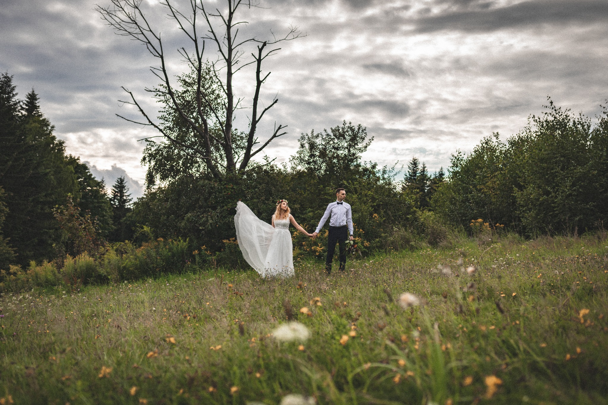 Para młoda trzymająca się za ręce spaceruje po łące, panna młoda w białej sukni z wiankiem na głowie, pan młody w koszuli z podwiniętymi rękawami i muchą, w tle las i pochmurne niebo.