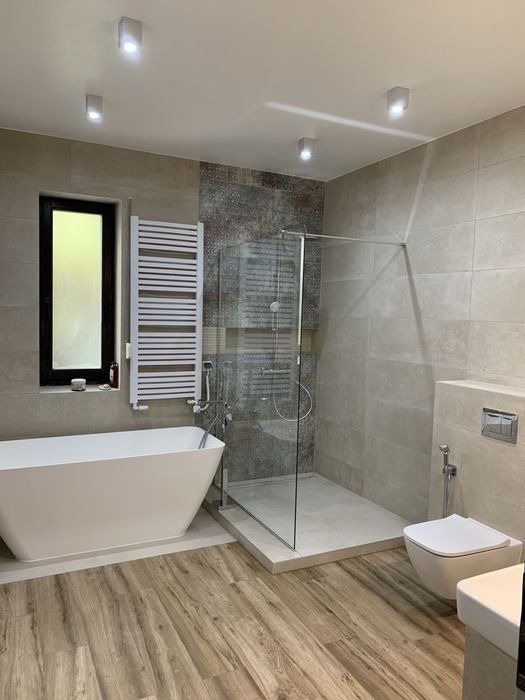 Nowoczesna łazienka z wolnostojącą wanną, kabiną prysznicową typu walk-in z odpływem liniowym, grzejnikiem drabinkowym i drewnopodobną podłogą, oświetlona punktowymi lampami sufitowymi.