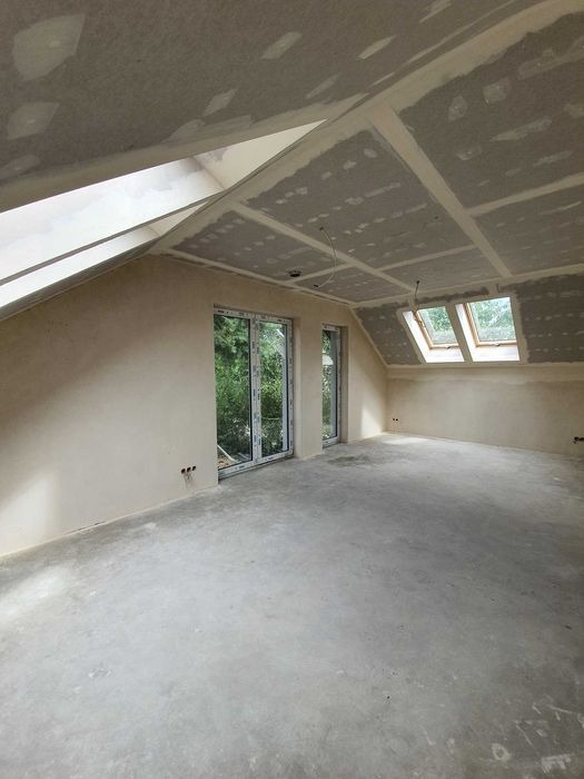 Poddasze w trakcie prac wykończeniowych: ściany po szpachlowaniu, widoczne łączenia płyt gipsowo-kartonowych na suficie, okna dachowe i balkonowe z widokiem na zieleń.